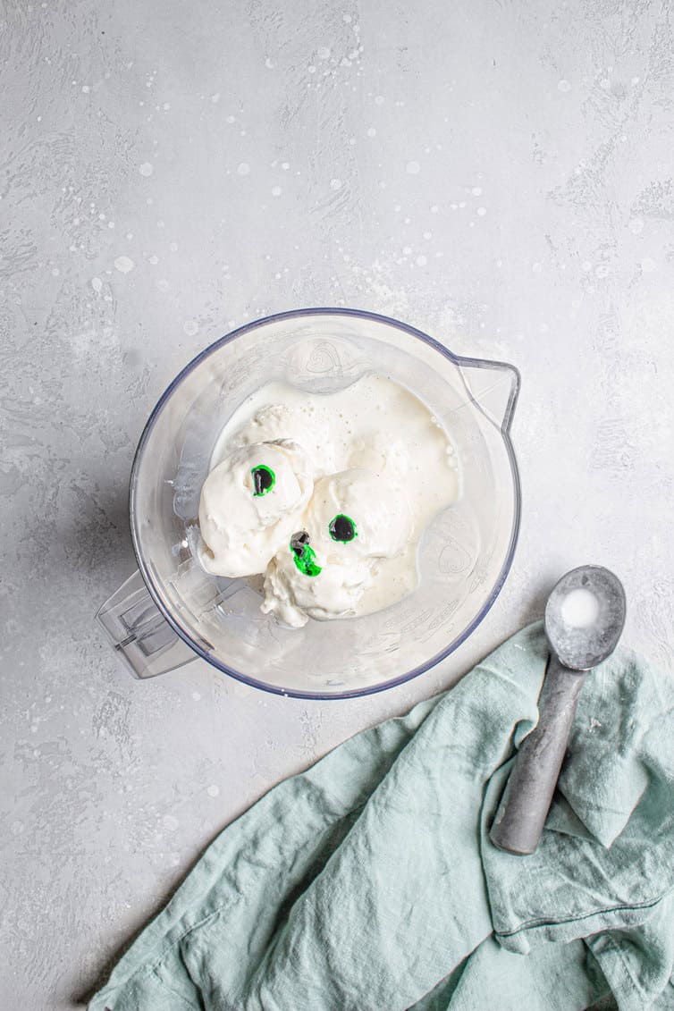 An overhead shot of a blender with vanilla ice cream and drops of green food coloring.