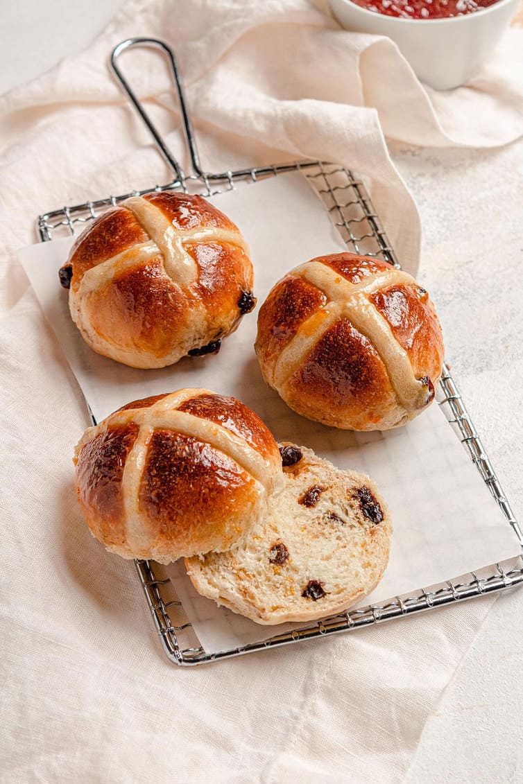 Three hot cross buns on a serving plate, one sliced in half.