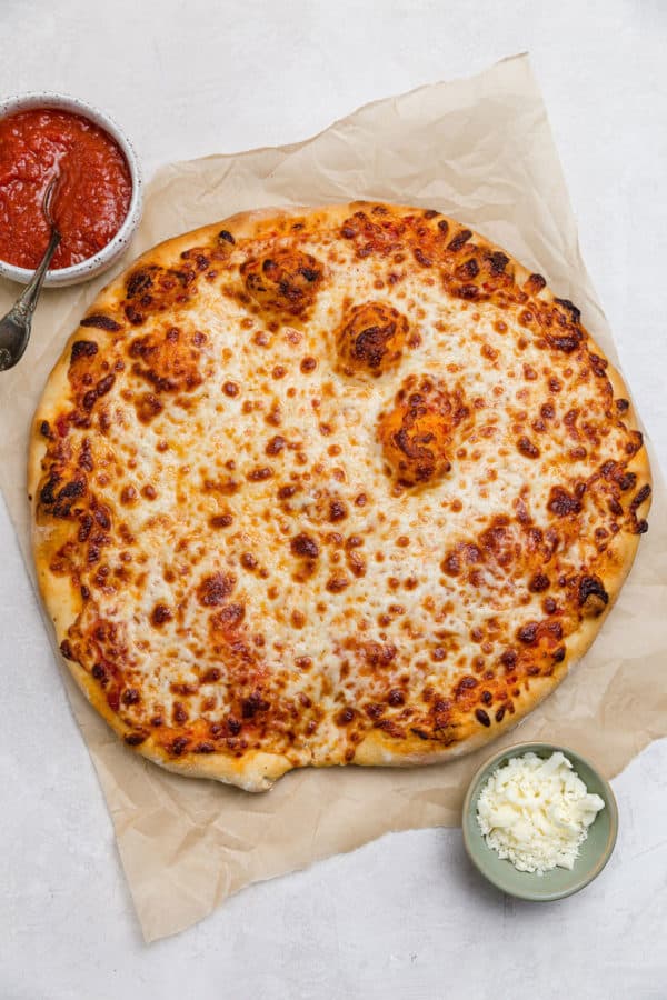 An overhead photos of a full cheese pizza with small bowls of sauce and cheese on the side.