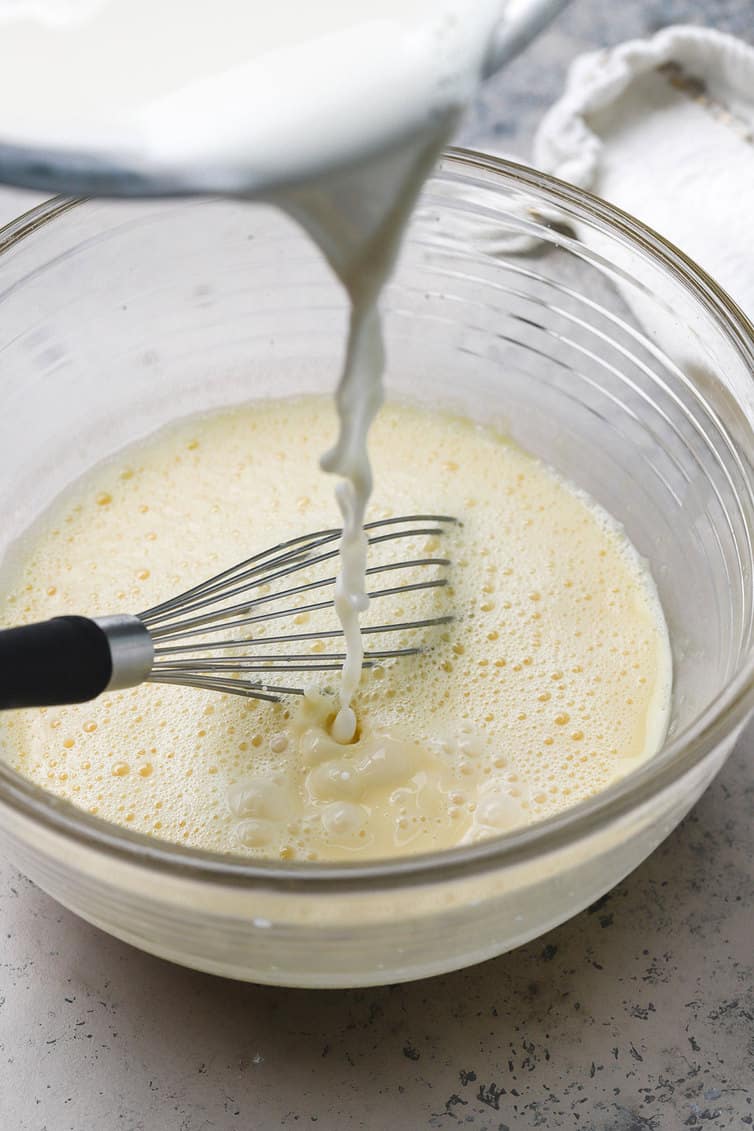 Warm cream mixture being whisked into beaten egg yolks.