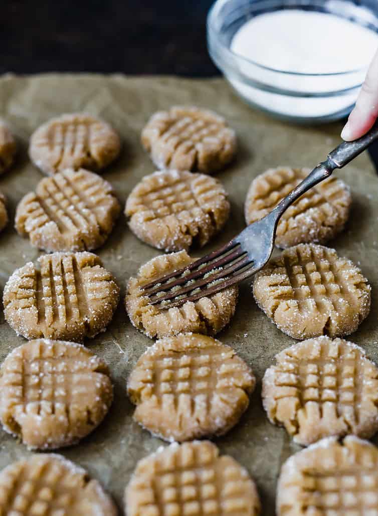 A fork making a crisscross pattern on the top of unbaked peanut butter cookies.
