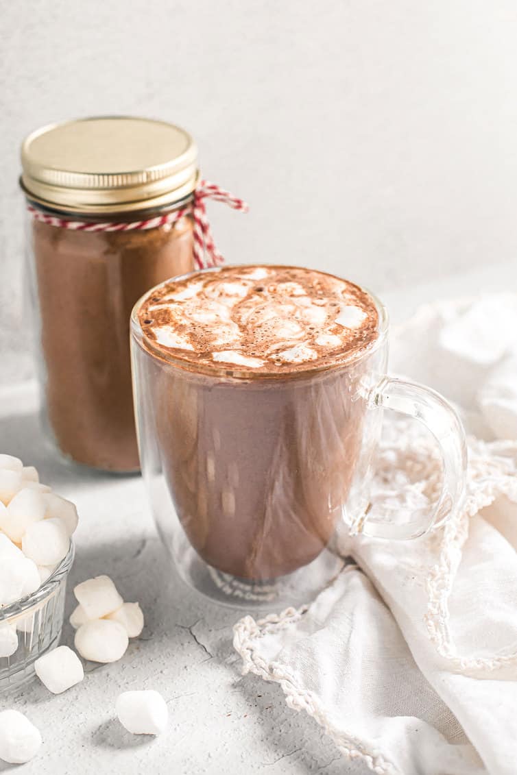 A cup of hot chocolate with marshmallows and a jar of hot chocolate mix in the background.