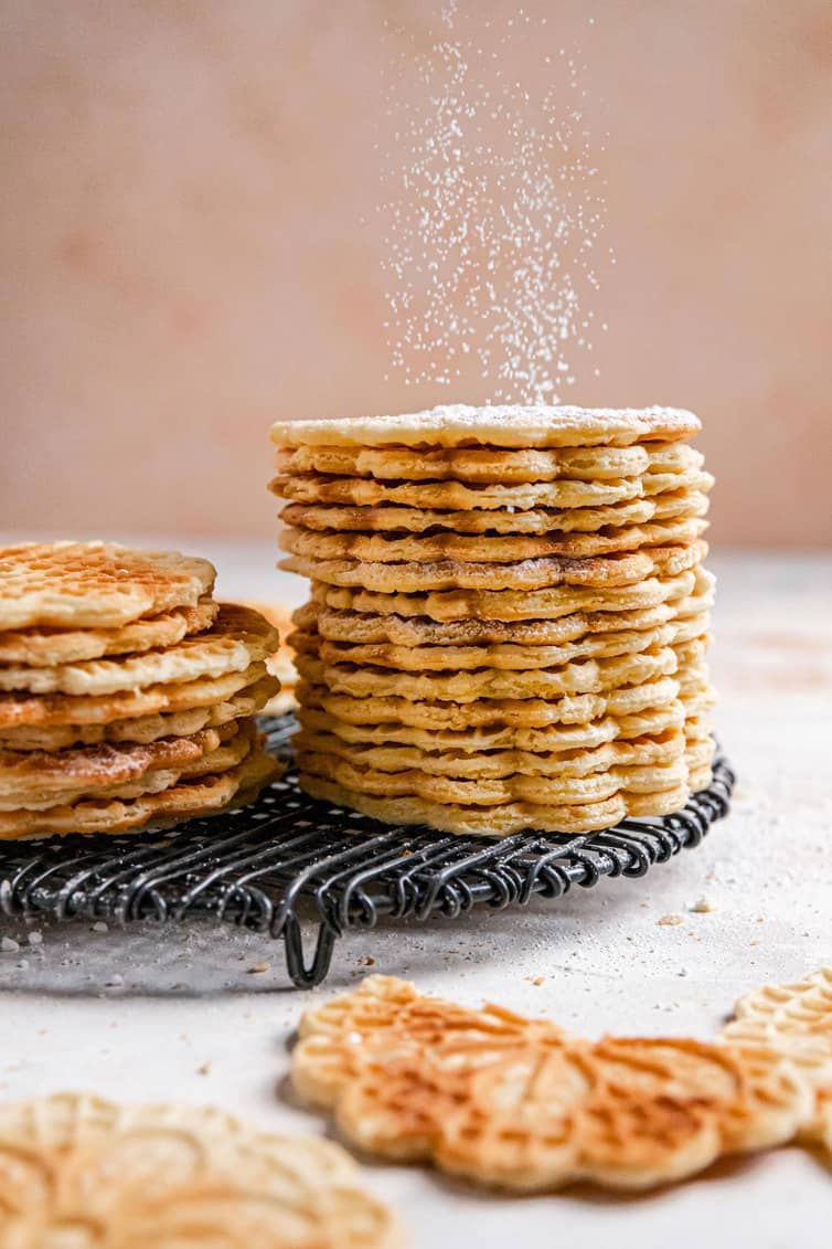 A stack of pizzelle on a cooling rack with powdered sugar being sprinkled on top.