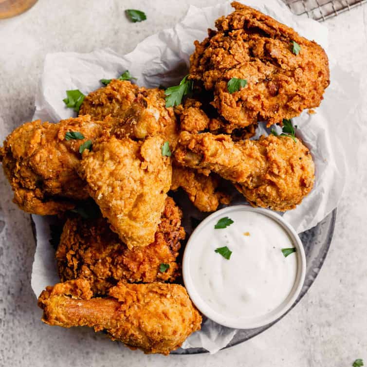 A silver plate with parchment paper topped with crispy fried chicken pieces with a small cup of ranch dressing.