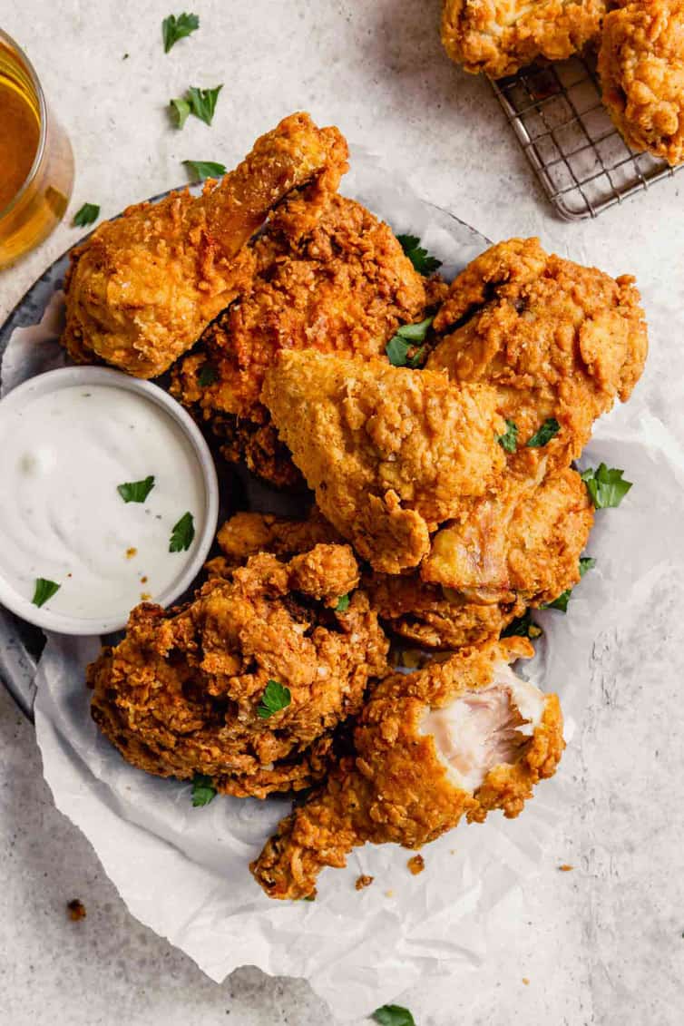 A fried drumstick on the right with a bite taken out at the bottom of a plate of buttermilk fried chicken with a small cup of ranch to the left.