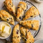 A circle of fresh buttermilk scones with blueberries on a black cooling rack covered in parchment paper with a small bowl of fresh sliced butter on the left side of the circle.