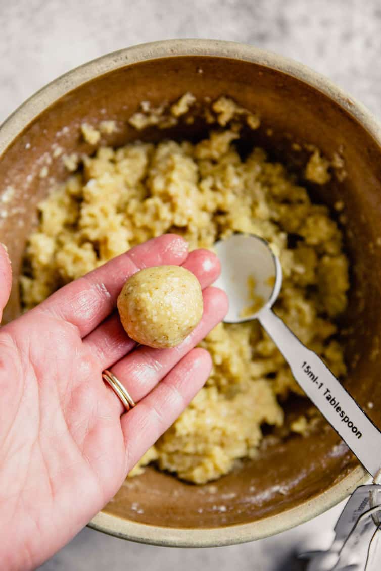 A hand holding 1-inch ball of matzo ball dough with a silver spoon in a bowl of matzo ball dough.