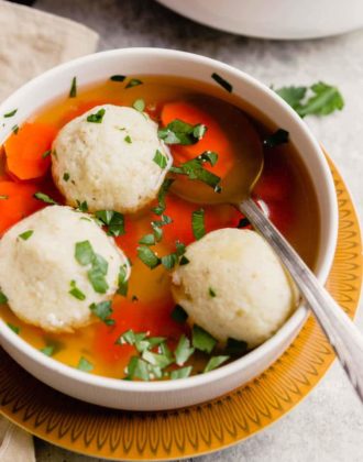 A warm bowl of matzo ball soup on a yellow plate with a silver spoon in the bowl topped with parsley.