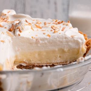 A square picture of fresh coconut pie in a glass pie plate showing the layers of graham cracker crust, coconut filling, and a whipped topping with toasted shredded coconut.
