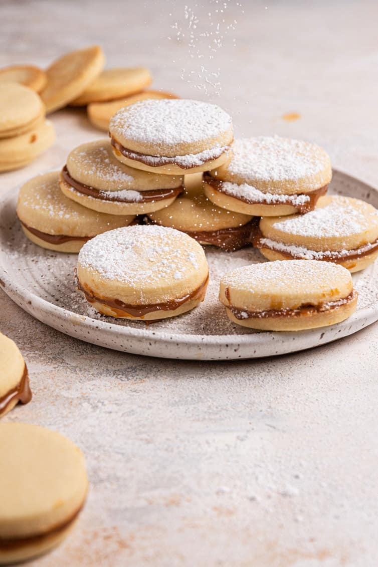 A white plate piled with alfajores dusted with powdered sugar.