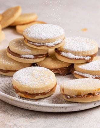 A square picture with a stack of alfajores on a white plate dusted with powdered sugar.