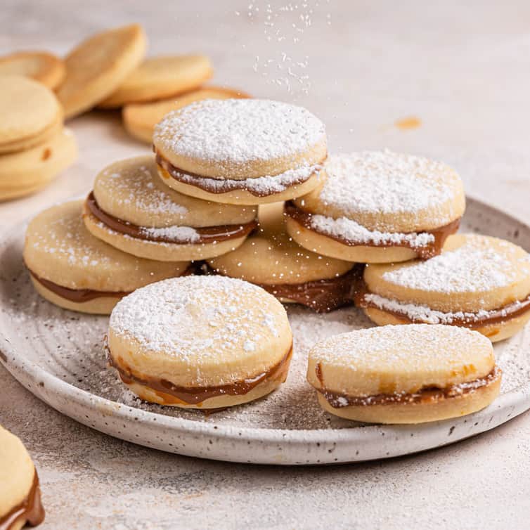 A square picture with a stack of alfajores on a white plate dusted with powdered sugar.