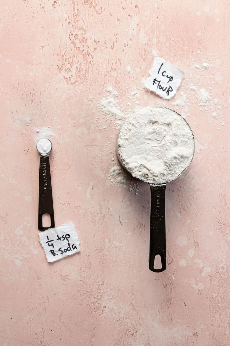 A measuring spoon with baking soda next to a measuring cup of flour.
