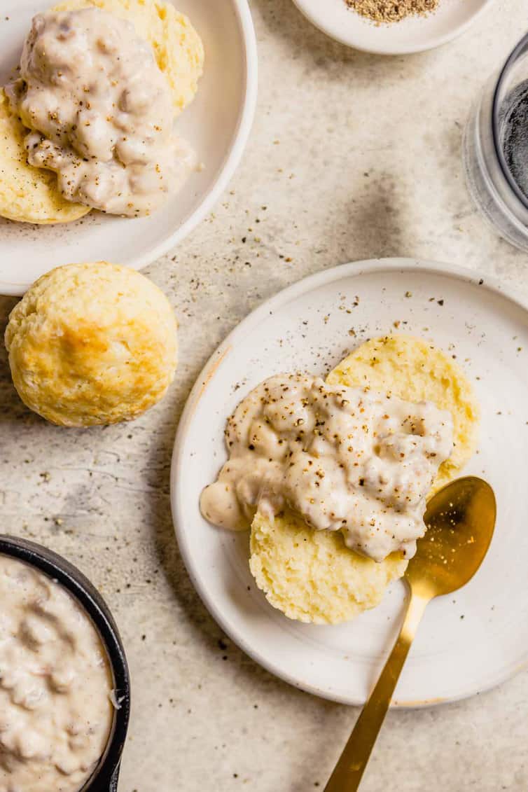 A plate of biscuits and gravy with a gold spoon on the right a biscuit to the left of the plate and another plate with biscuits and gravy in the top left.