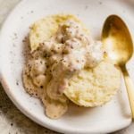 A biscuit topped with sausage gravy on a white plate with a gold spoon on the right.