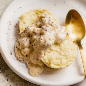 A biscuit topped with sausage gravy on a white plate with a gold spoon on the right.