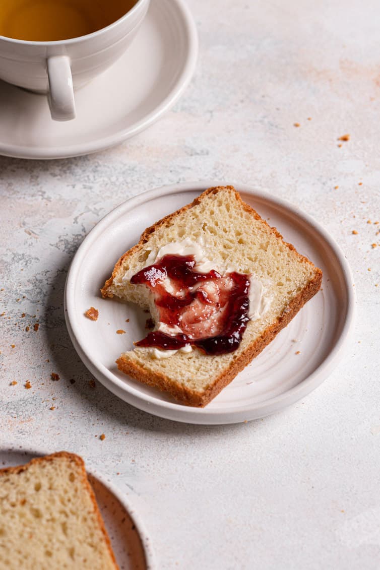 A slice of English muffin bread on a white plate spread with butter and jam.