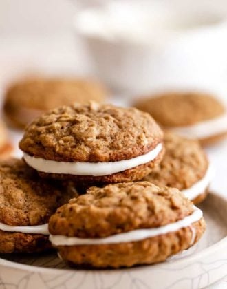 A stack of oatmeal cream pies on a white plate with a white bowl in the back right.