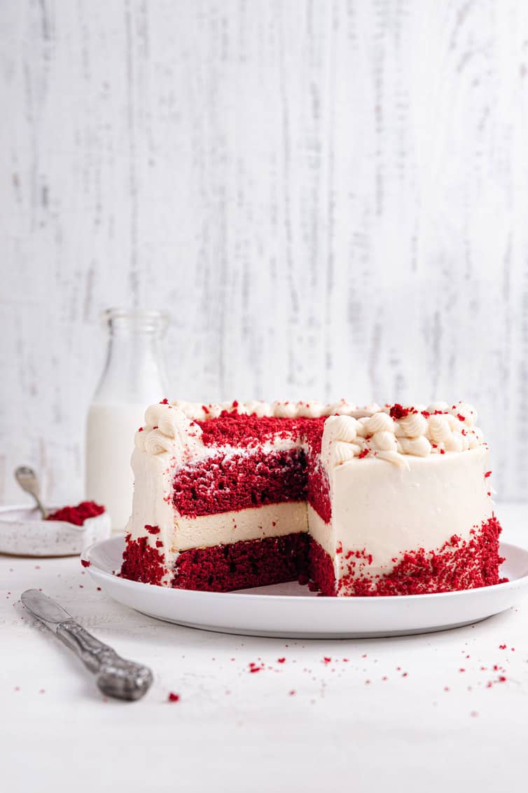 A large red velvet cheesecake on a white plate in front of a jug of milk.