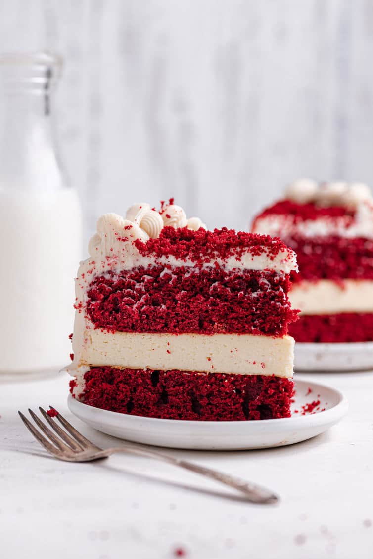 A slice of red velvet cheesecake on a white plate from the side with a fork in front.