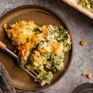 A brown plate with a scoop of broccoli casserole with a fork with a wooden handle on a grey counter.