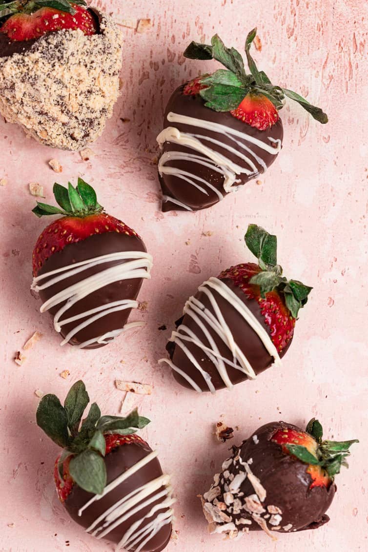 Six chocolate strawberries topped with chocolate and nuts on a pink counter.