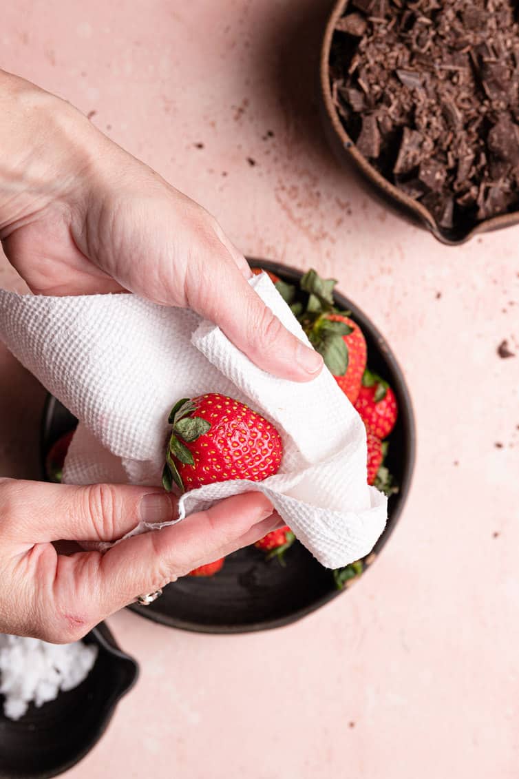 A hand holding a paper towel drying a strawberry over a bowl of fresh strawberries.