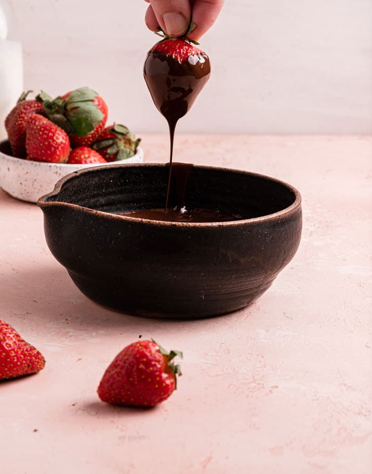 A brown bowl with melted chocolate and a hand holding a strawberry freshly dipped in chocolate drizzling back in the bowl.
