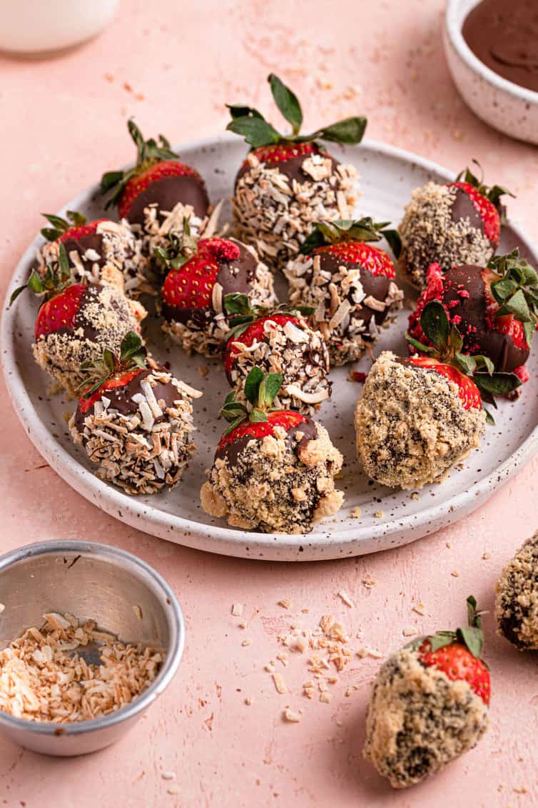 A white plate of chocolate strawberries topped with nuts with chocolate in the back and a strawberry on the counter in the front right.