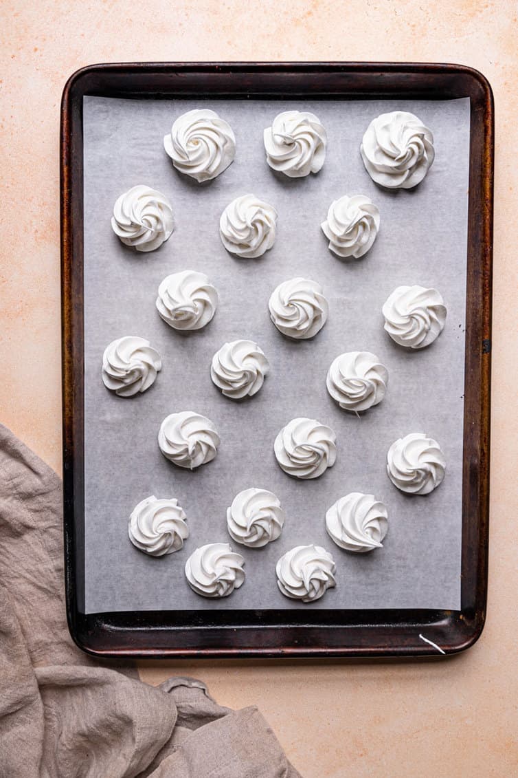 A parchment paper lined baking sheet with piped meringue cookies on a pink counter.