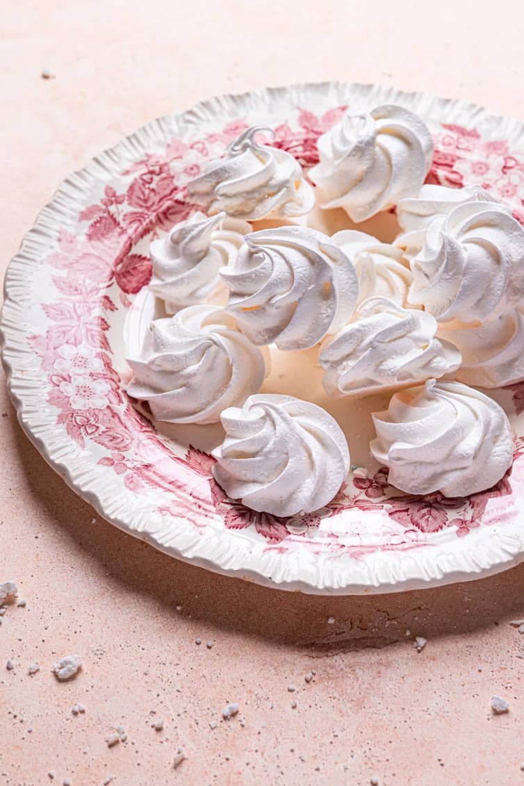 A from the side photo of a pink and white plate with white meringue cookies on a pink counter.