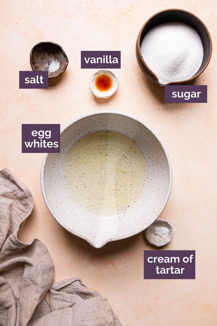 Ingredients for meringue cookies in bowls, labeled with purple labels, on a pink counter.