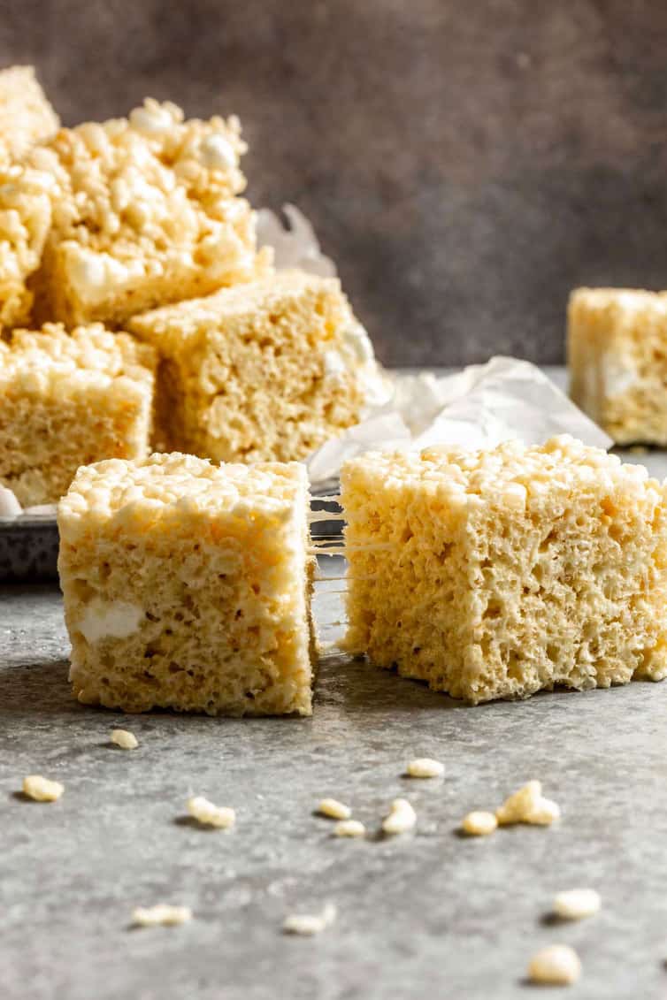 From the front, two tall rice krispie treat squares in front of a pile of rice krispie treats on a parchment paper lined plate with rice krispies in front.