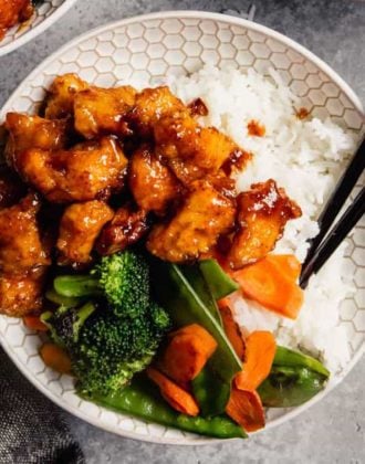 A white bowl of baked sweet and sour chicken with broccoli, rice, and chopsticks.