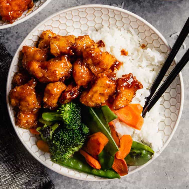 A white bowl of baked sweet and sour chicken with broccoli, rice, and chopsticks.