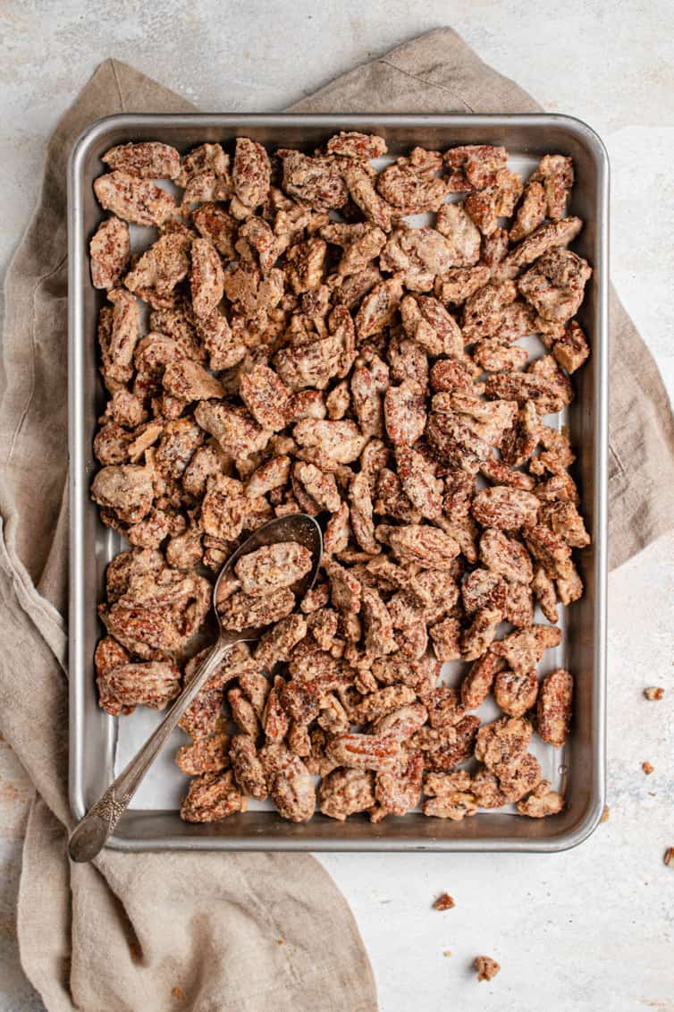 A metal baking sheet with candied pecans on a light brown towel on a white counter.