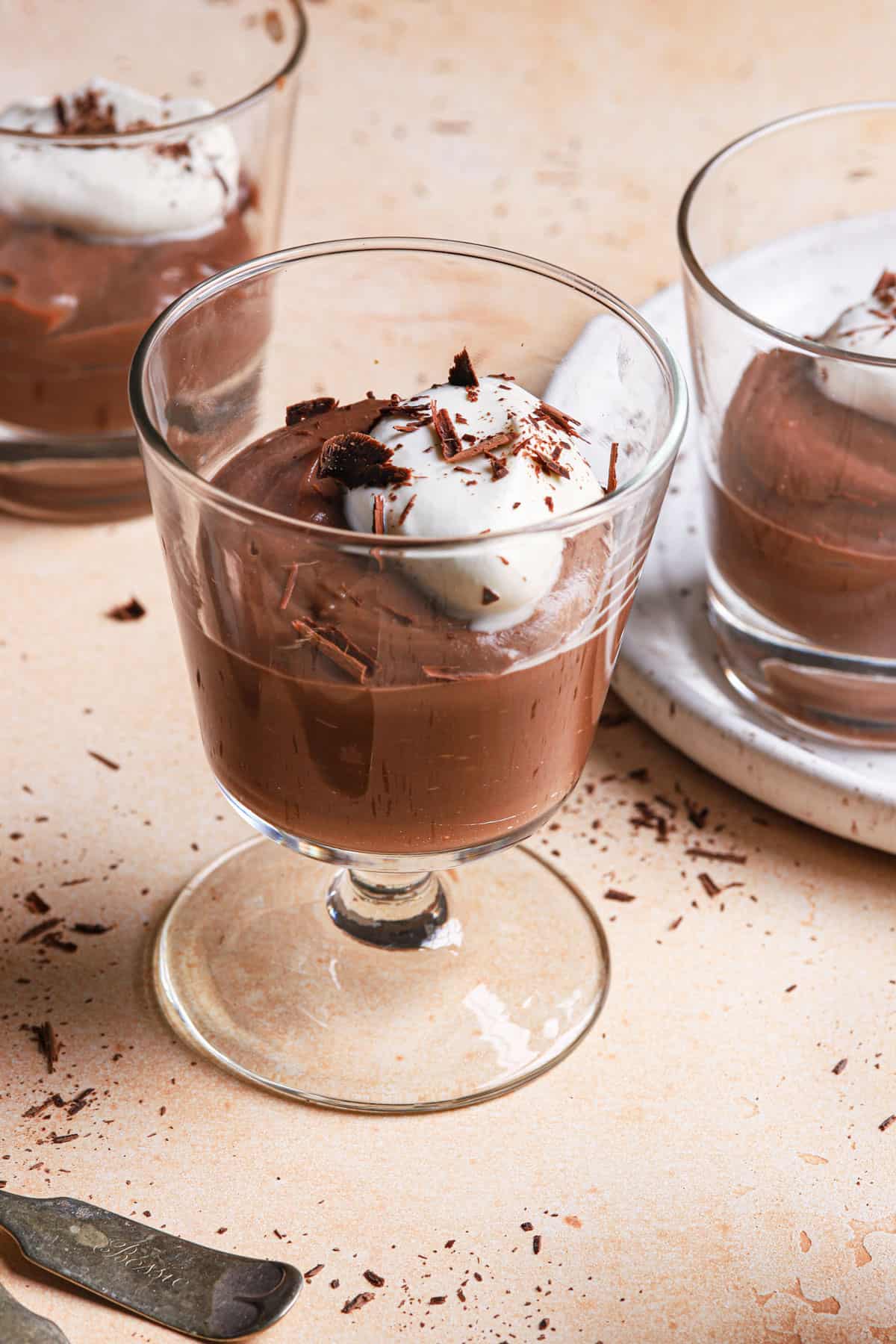 A close up of a glass of chocolate pudding with whipped cream and chocolate shavings with two cups behind it.