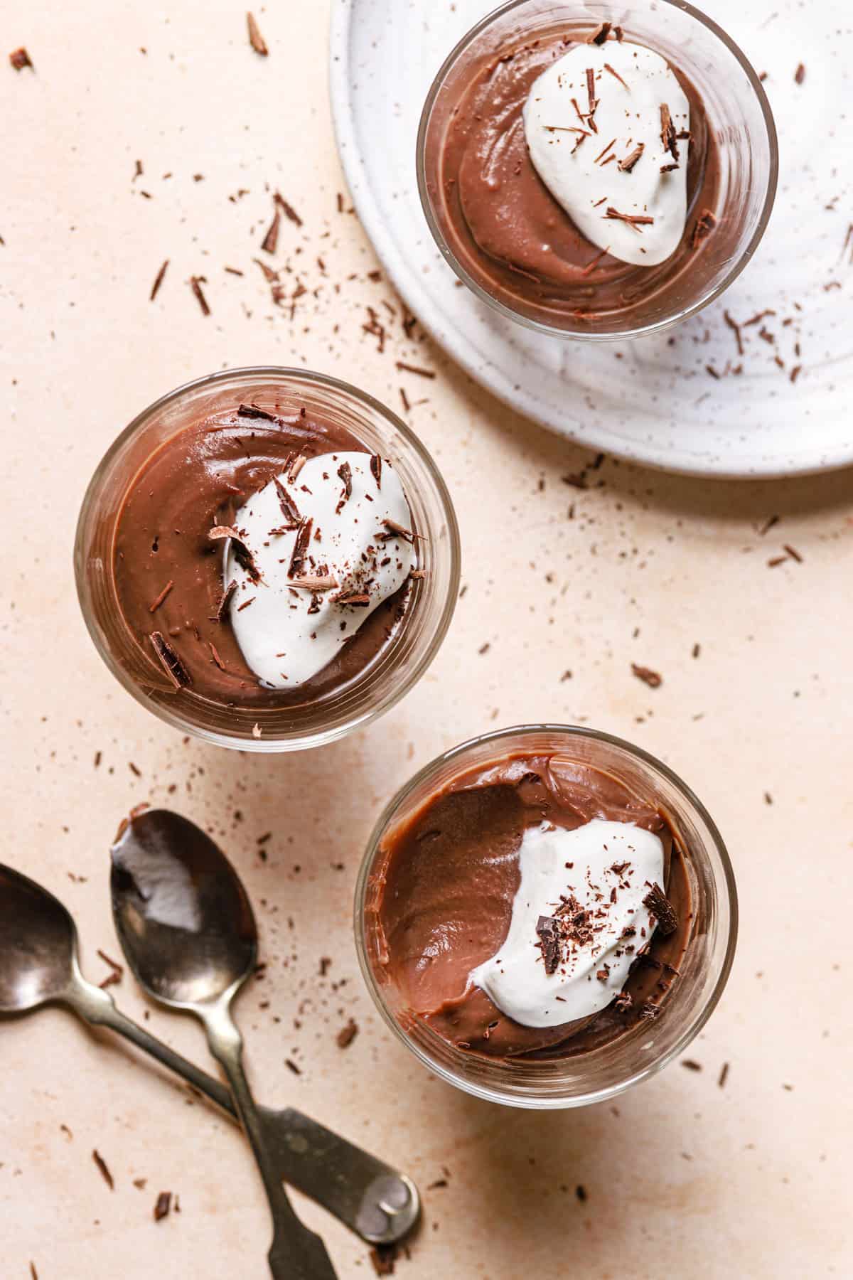 A top down photo with two glasses of chocolate pudding topped with whipped cream on the pink counter and the bottom glass has a bite taken out with two spoons to the left and a white plate with a glass of chocolate pudding on top with whipped cream and chocolate shavings.