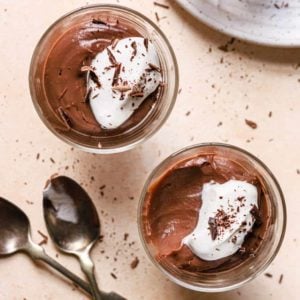 A square photo of two glasses of chocolate pudding with a scoop taken out of the bottom right one.