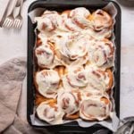 A baking pan with cinnamon rolls covered in cream cheese frosting with a stack of forks in the top left.