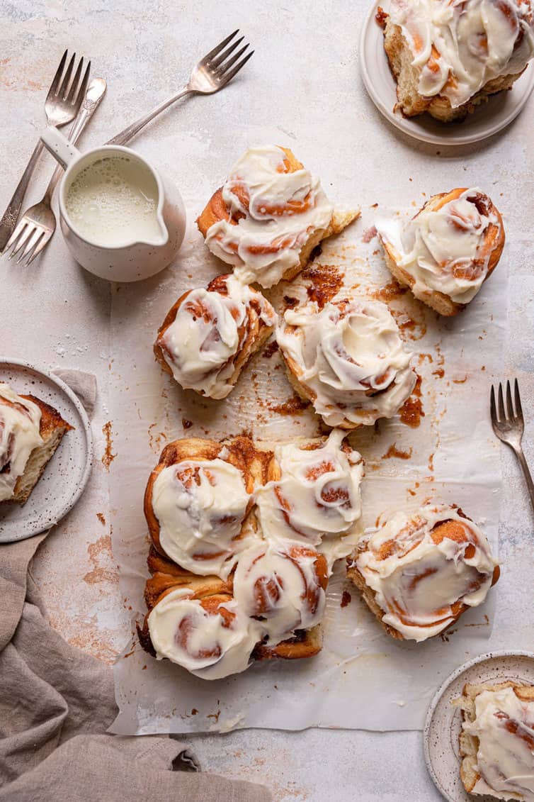 Cinnamon rolls spread out on a piece of parchment paper with other cinnamon rolls on plates in the top and bottom right corner and middle left side.