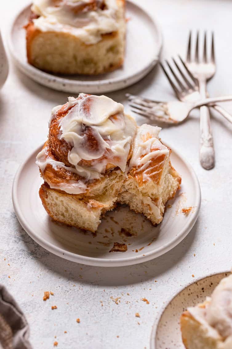 A cinnamon roll on a plate with a bite taken out and a cinnamon roll on a plate in the back with forks stacked.