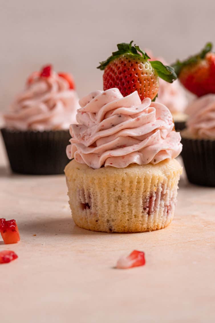 A fresh strawberry cupcake topped with strawberry frosting without a wrapper on a pink counter with a piece of chopped strawberry.