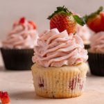 A square photo of a strawberry cupcake without a wrapper topped with strawberry meringue buttercream and a fresh strawberry.