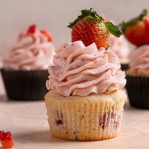 A square photo of a strawberry cupcake without a wrapper topped with strawberry meringue buttercream and a fresh strawberry.