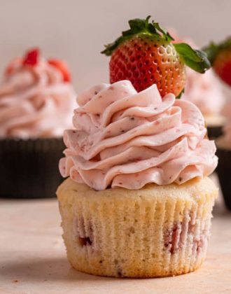 A square photo of a strawberry cupcake without a wrapper topped with strawberry meringue buttercream and a fresh strawberry.