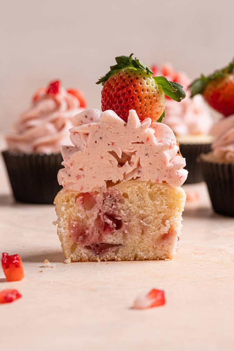 A sliced cupcake showing the inside of the strawberry cupcake and frosting topped with a strawberry.