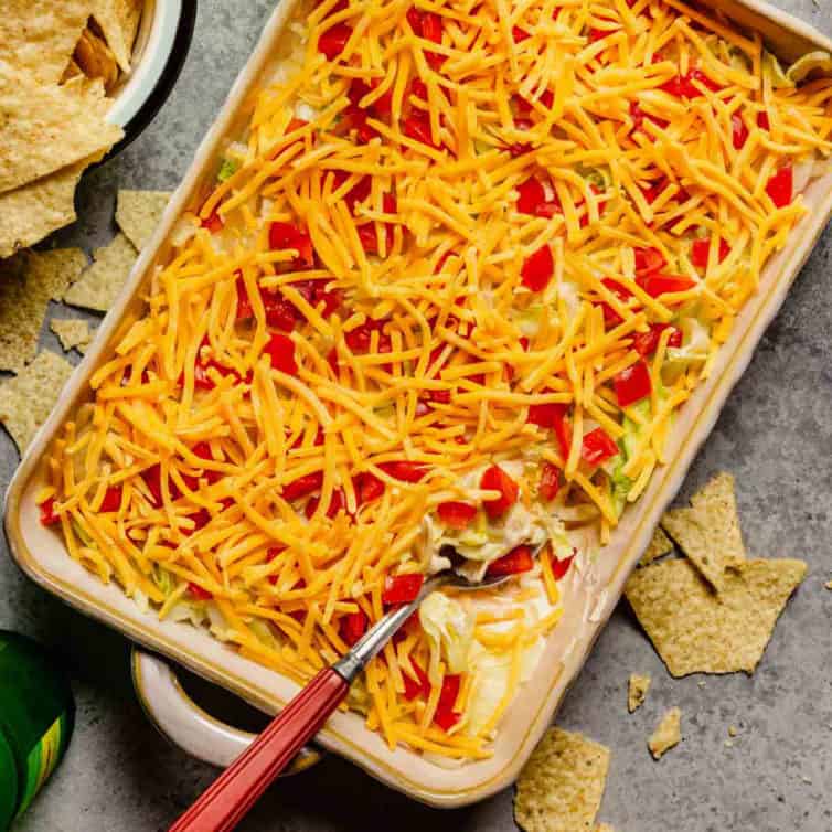 A square photo of taco dip with a spoon and a bowl of tortilla chips to the left.