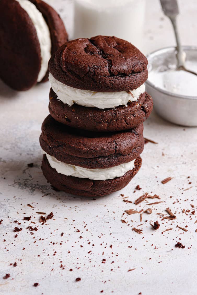 A white counter with chocolate shavings and two whoopie pies stacked on top of each other with a bowl of marshmallow fluff filling in the back right.