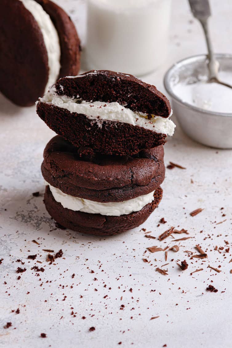 Two whoopie pies stacked with the filling exposed on the top and a glass of milk in the back with a bowl of marshmallow filling in the back right.
